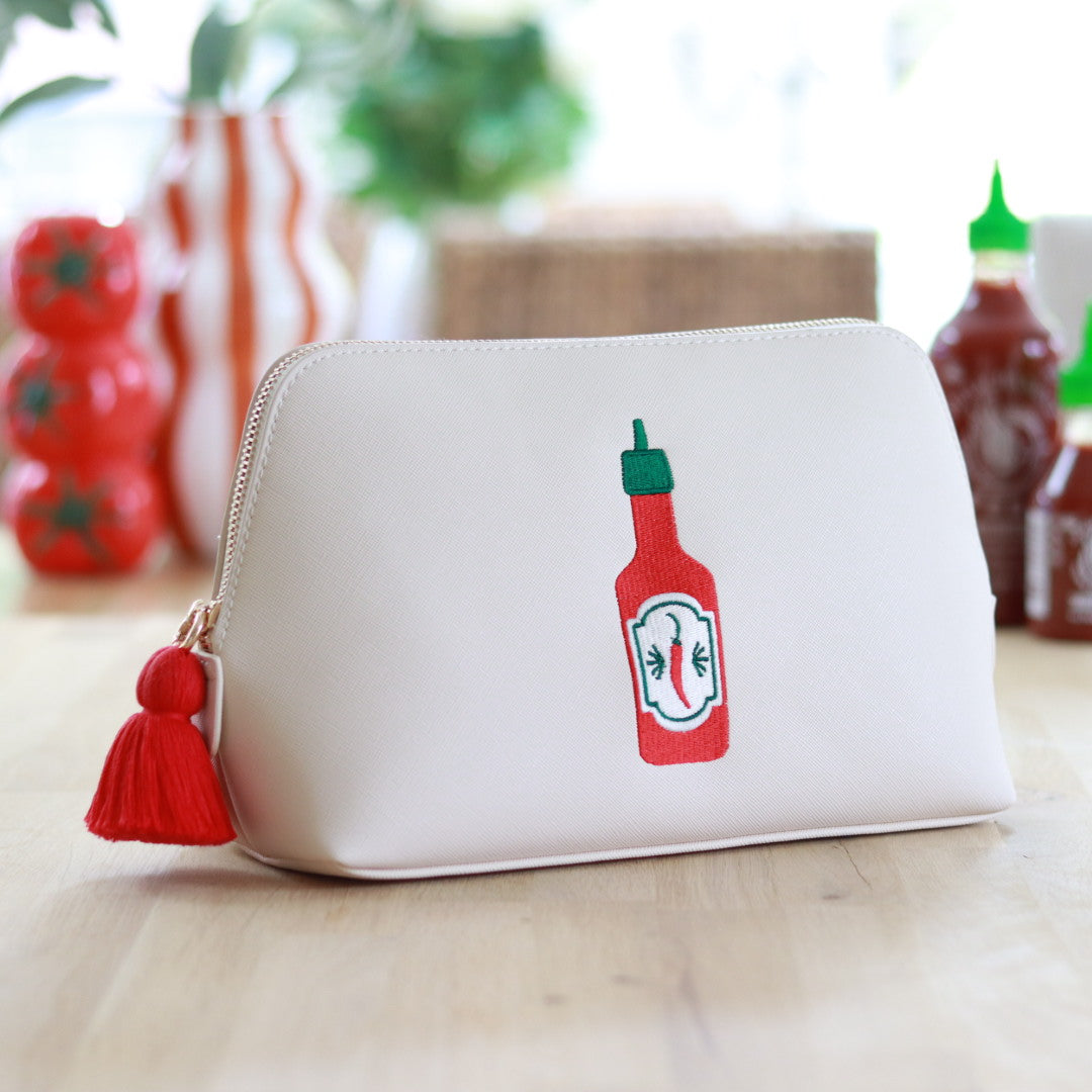 Cream faux saffiano leather zippered pouch with a red tassel zipper and an embroidered red and green hot sauce bottle design on the front, displayed on a wooden surface with colorful kitchen décor in the background.