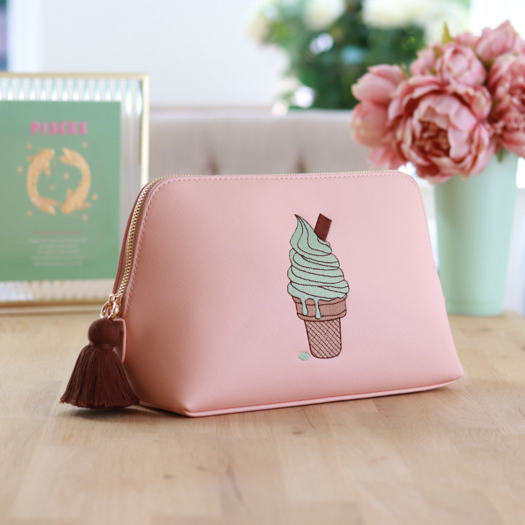 Light pink faux saffiano leather zippered pouch with a brown tassel zipper and an embroidered mint, beige & brown mint flavoured ice cream cone with chocolate flake on the front, displayed on a wooden surface with colorful décor in the background.