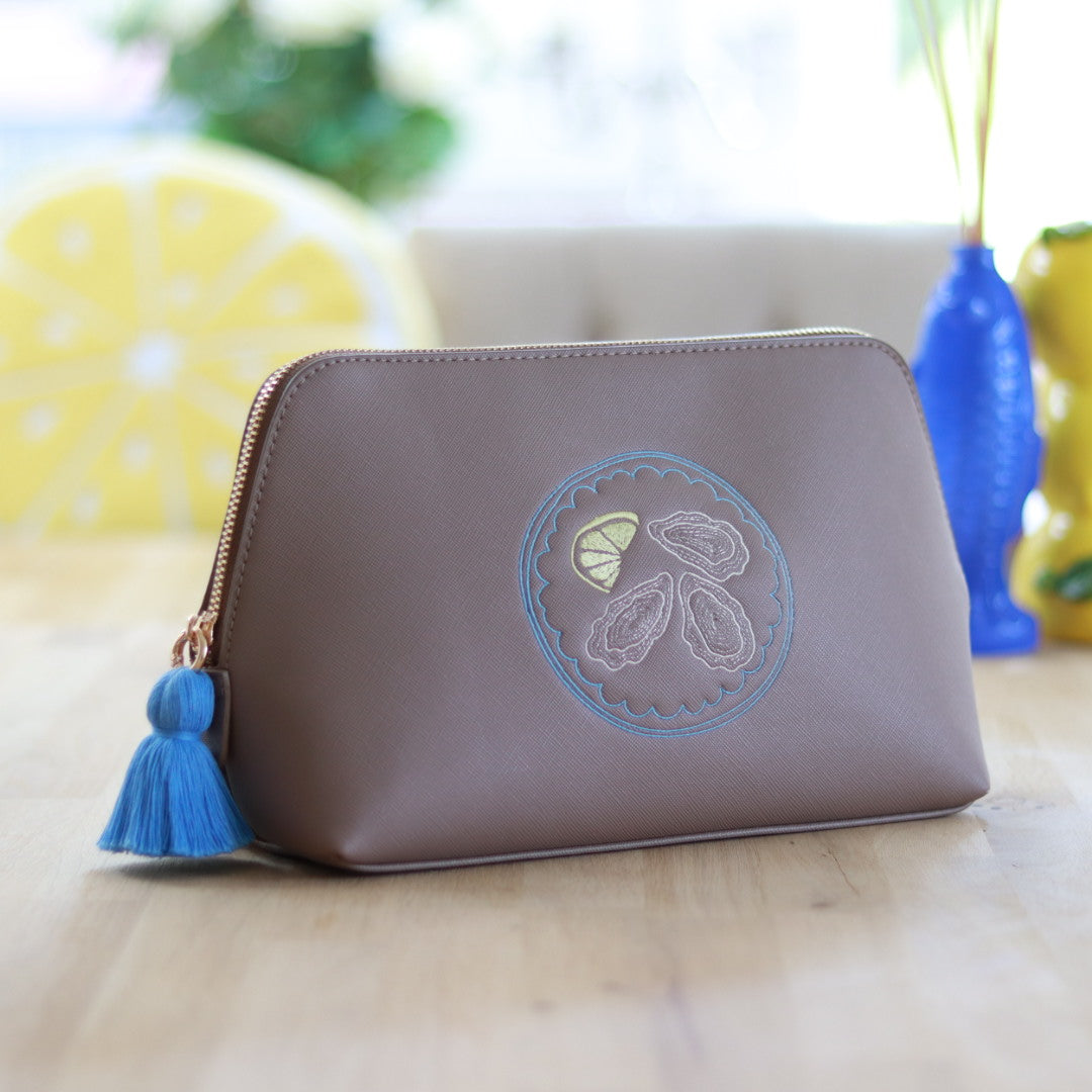 Taupe zippered pouch with a blue tassel zipper and embroidered oyster and lemon slice design in cream, blue & yellow, displayed on a wooden surface with colorful decor in the background.