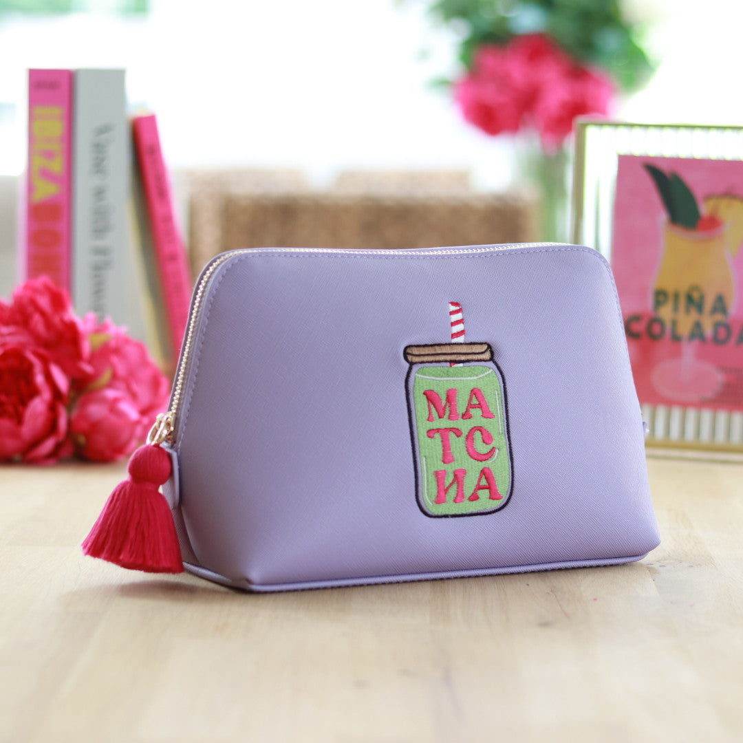 Lilac zippered pouch with a hot pink tassel zipper and embroidered matcha jar design in green and hot pink, displayed on a wooden surface with colorful books and floral decor in the background.